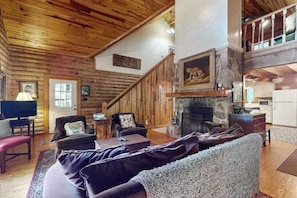 A log cabin interior with a fireplace, wooden walls, a staircase, and a dining area.