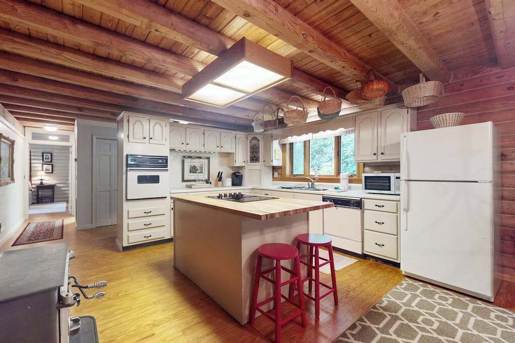 A kitchen with wooden beams, white cabinets, a central island, red stools, and a refrigerator.