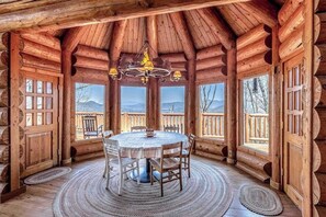 A wooden cabin interior with a round dining table, chairs, a chandelier, and large windows offering mountain views. - Mountain-view log home with hot tub, firepits, pool table & air hockey (Franklin)