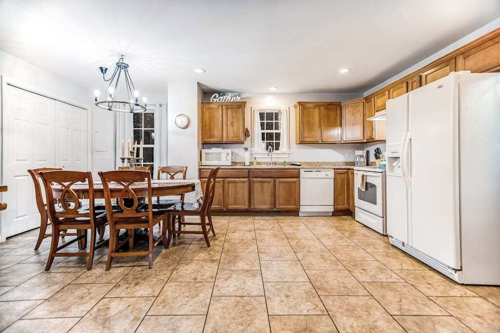 A kitchen with wooden cabinets, a dining table with chairs, a chandelier, and a clock on the wall.