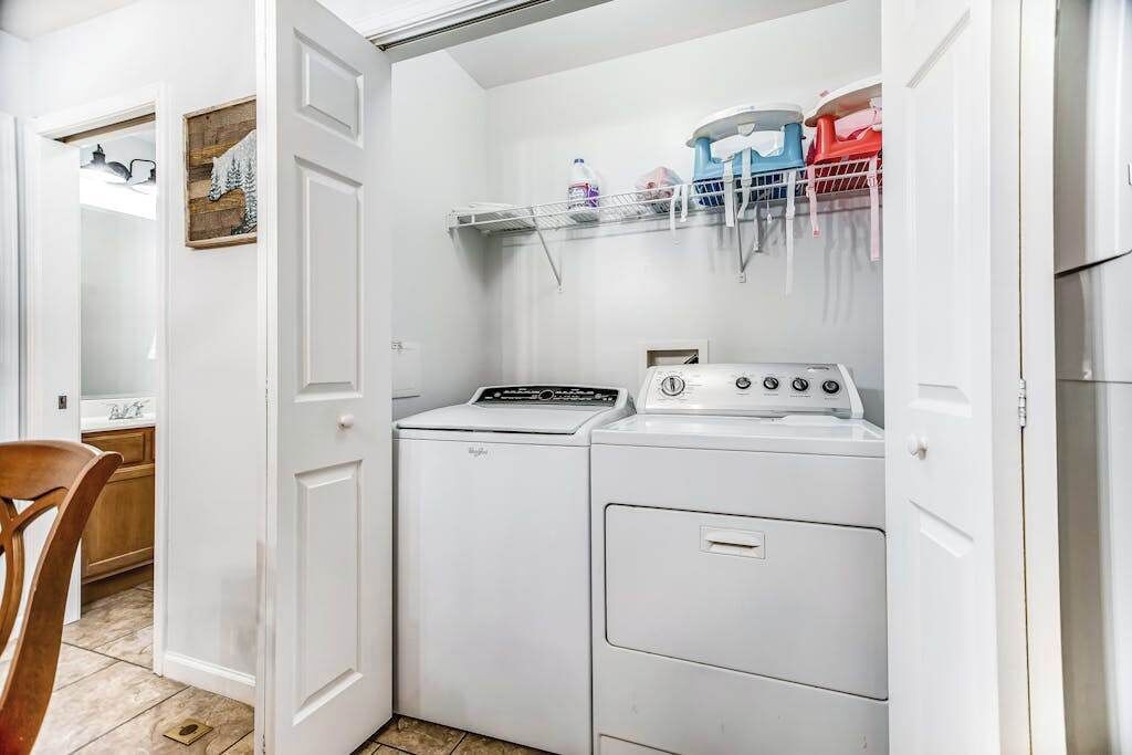 A laundry room with a washing machine and dryer, a shelf with cleaning supplies, and a door leading to another room.