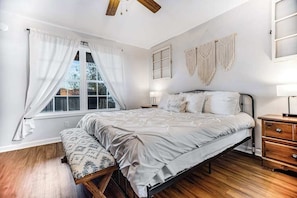 Ein Schlafzimmer mit Bett, Nachttisch, Bank, Fenster mit Vorhängen und Wanddekoration.