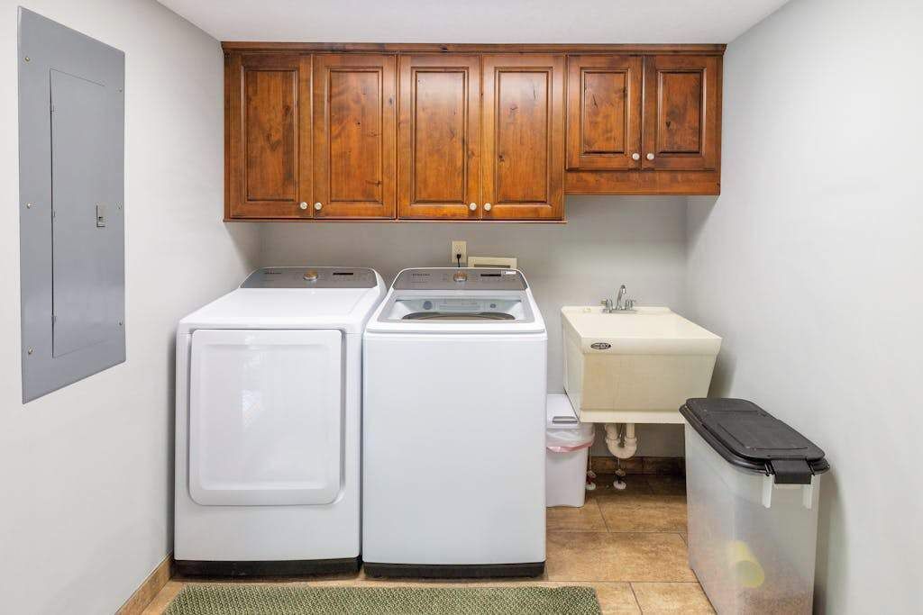 A compact laundry room with two white washing machines, a sink, wooden cabinets, and a trash bin.