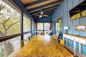 Ein Essbereich mit Holztisch, Stühlen, Deckenventilator und Blick auf Bäume durch große Fenster.