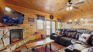 A cabin interior with a stone fireplace, leather sofa, television, ceiling fan, and wooden walls.