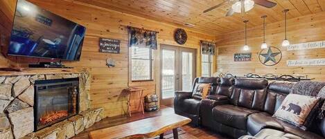 A cabin interior with a stone fireplace, leather sofa, television, ceiling fan, and wooden walls.