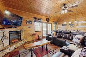 Ein Ferienhaus-Innenraum mit Kamin aus Stein, einer Ledersofa, Fernseher, Deckenventilator und Holzwänden.