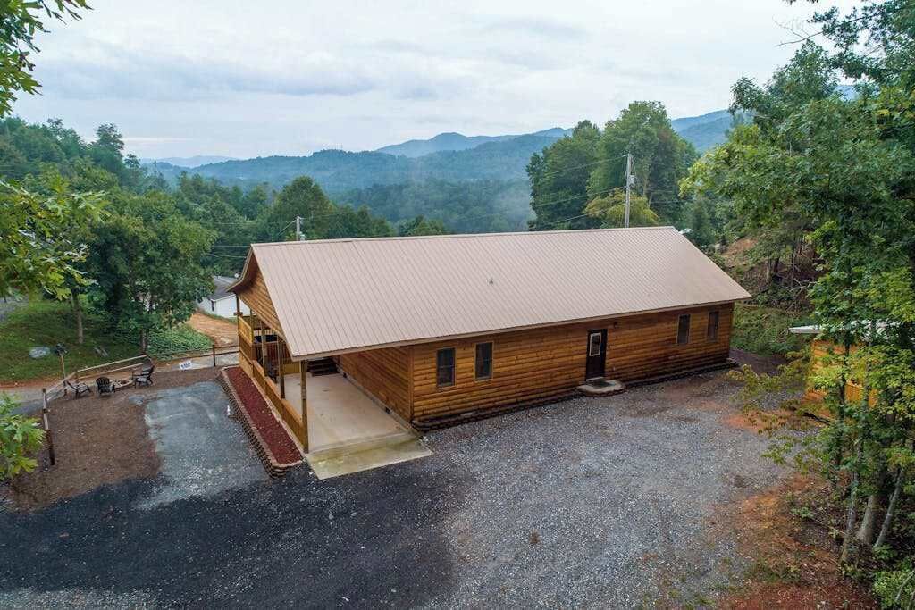 A wooden cabin with a metal roof, surrounded by a gravel driveway and a wooden deck, set against a backdrop of trees and mountains.