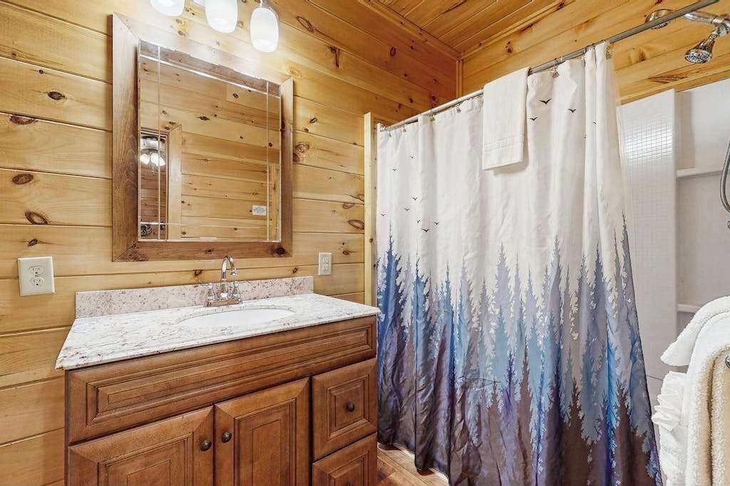 Baño de cabaña de madera con tocador de madera, un lavamanos, un espejo y una cortina de ducha con diseño de montaña.