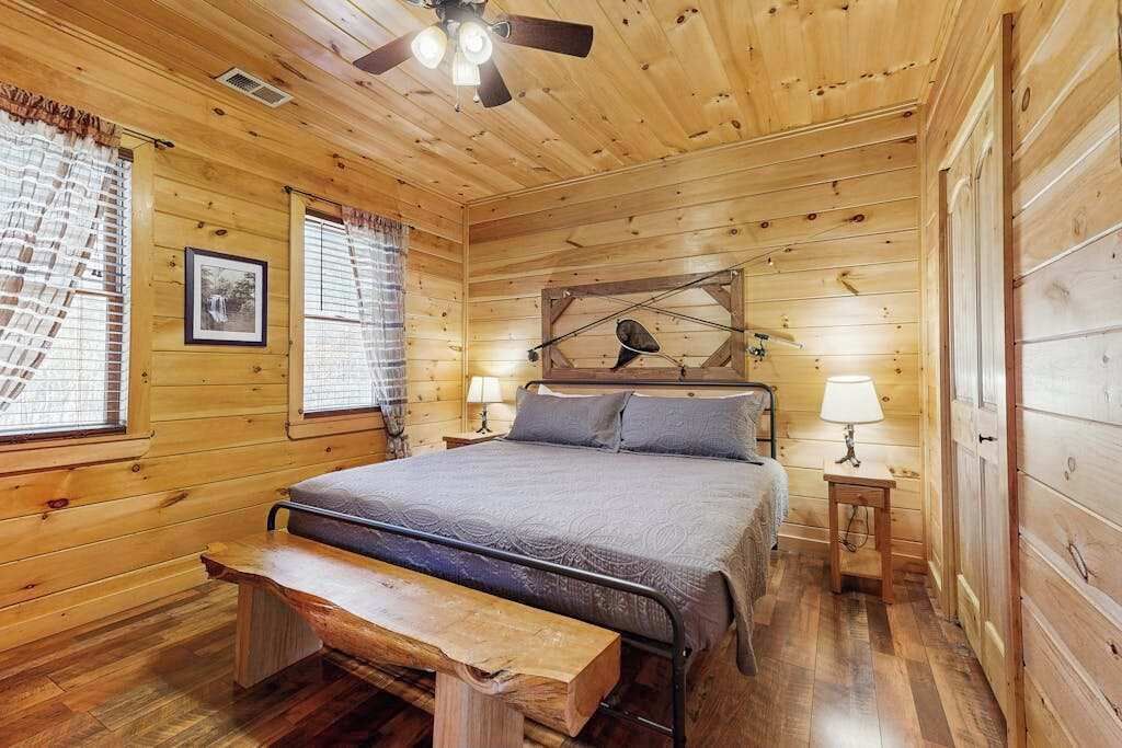 A wooden cabin bedroom with a bed, bench, two bedside tables, a ceiling fan, and a window with curtains.