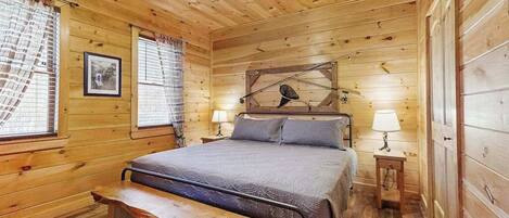 A wooden cabin bedroom with a bed, bench, two bedside tables, a ceiling fan, and a window with curtains.