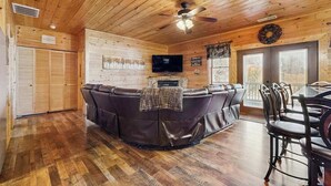 A wooden cabin interior with a leather couch, dining table, chairs, a ceiling fan, and a television.