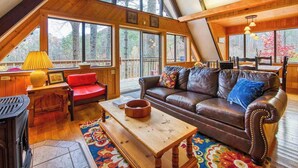 A cozy loft-style room with a wooden ceiling, large windows, a leather sofa, a red chair, and a wooden table. - Chalet at Peek's Creek (Franklin)