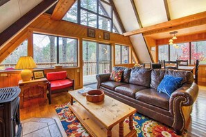 Ein gemütliches Loft-Zimmer mit Holzbalkendecke, großen Fenstern, einem Ledersofa, einem roten Sessel und einem Holztisch.