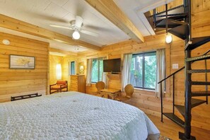 Eine Holzhütte mit einem Bett, einem Schreibtisch, einem Stuhl, einem Fernseher und einer Treppe.