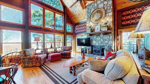 A spacious living room with a stone fireplace, large windows, and a ceiling fan.