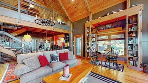 A spacious loft with a high ceiling, exposed wooden beams, a large sofa, a dining area, and a bookshelf.