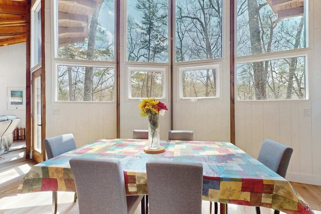 Un coin repas avec une table recouverte d’une courtepointe, entourée de chaises, et de grandes fenêtres donnant sur la forêt.