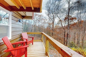 Eine Holzterrasse mit zwei roten Adirondack-Stühlen, einem Fenster und Blick auf einen Wald.