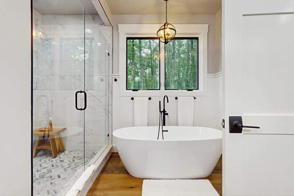 A modern bathroom with a freestanding bathtub, a glass shower enclosure, and a window with a view of trees.