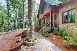 A stone house with a porch, a staircase, and a forested backdrop. - The Fox Den (Cashiers)