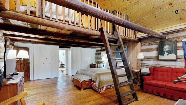 A rustic cabin interior with a loft bedroom, wooden floors, a ladder, a red sofa, and a painting on the wall. - Crackerbox Palace (Sylva)