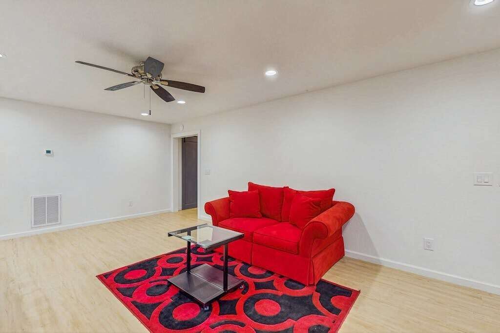 Una habitación con ventilador de techo, un sofá rojo, una mesa de centro de vidrio y una alfombra estampada.