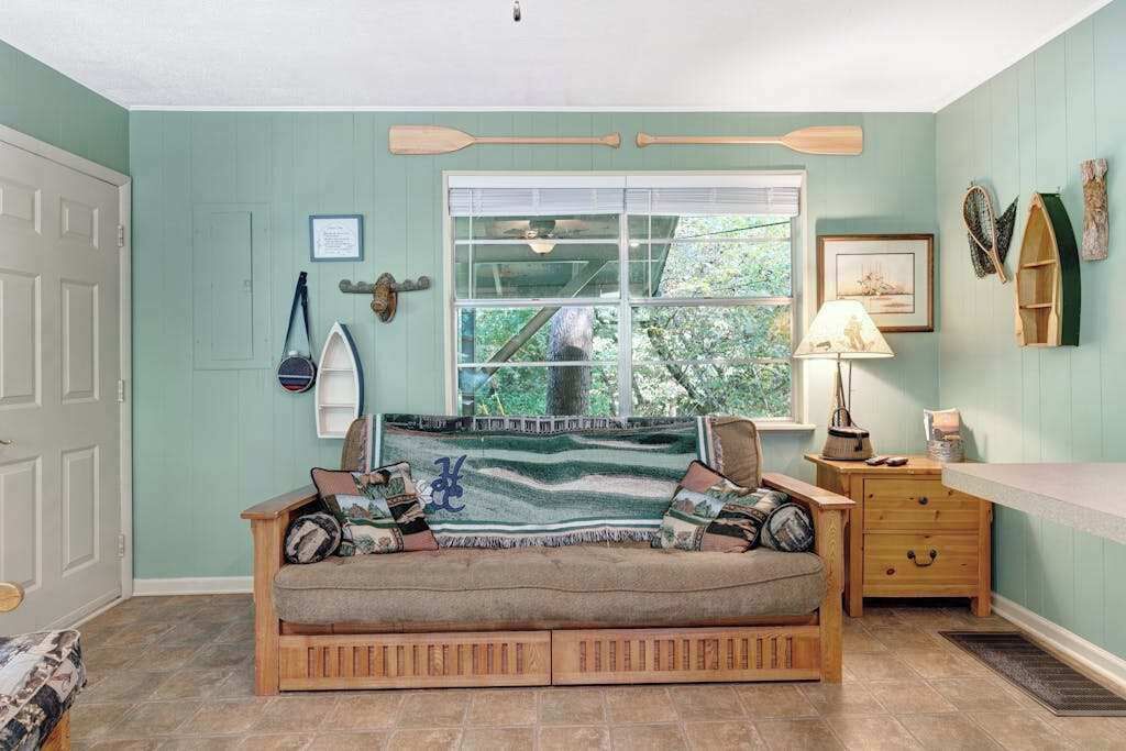 A cozy room with a wooden sofa, a window with a view of greenery, a small wooden side table, and a wall-mounted shelf with decorative items.