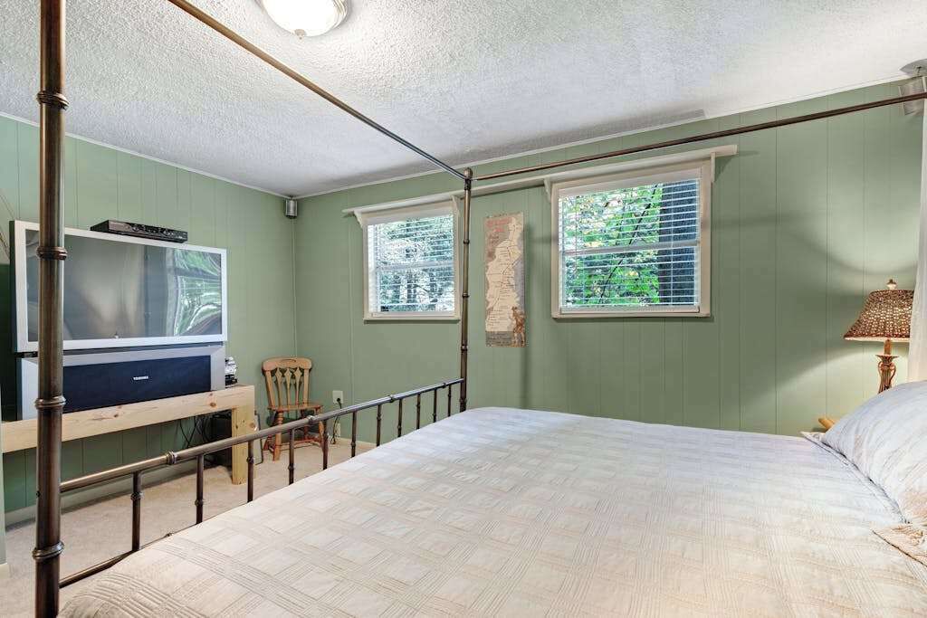 A bedroom with a bed, a television, a chair, and two windows with blinds.