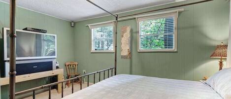 A bedroom with a bed, a television, a chair, and two windows with blinds.