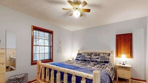 Une chambre avec un lit en bois, un ventilateur de plafond, une fenêtre avec des stores, une table de chevet avec une lampe et un meuble fixé au mur.