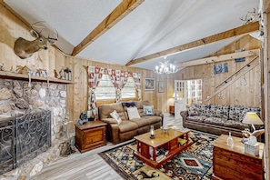 Un intérieur rustique avec des murs en bois, une cheminée en pierre, deux canapés, un lustre et un escalier.