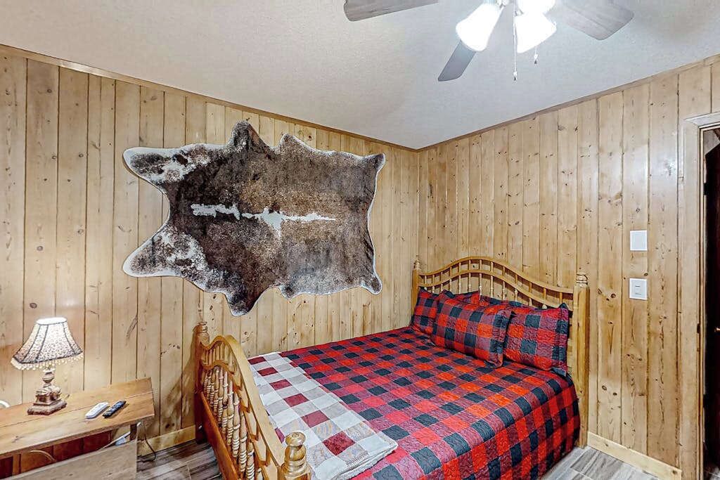 A room with wooden walls, a bed with a red and black checkered bedspread, a wooden headboard, a nightstand with a lamp, and a mounted animal hide on the wall.
