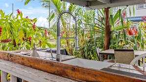 Une terrasse en bois avec un évier en métal, entourée d’une végétation tropicale luxuriante et dotée d’un espace couvert avec des chaises et une table.