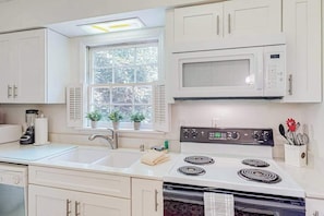 A kitchen with white cabinets, a microwave, a stove, a sink, and a window with plants.