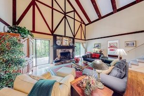 A spacious living room with a fireplace, wooden beams, and a large window overlooking a deck.
