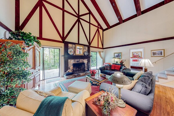 A spacious living room with a fireplace, wooden beams, and a large window overlooking a deck.