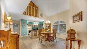 Spaziosa sala da pranzo con soffitto alto e travi in legno, una cucina sullo sfondo e una casetta da uccelli decorativa su un tavolo.