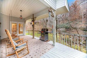 Eine überdachte Veranda mit Schaukelstühlen, einem Grill und Blick auf einen bewaldeten Bereich.
