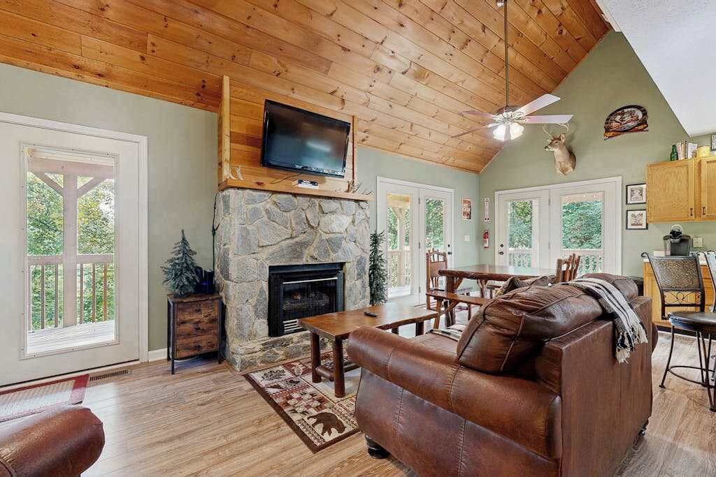 A cozy living room with a stone fireplace, leather sofa, wooden dining table, and a television mounted above the fireplace.