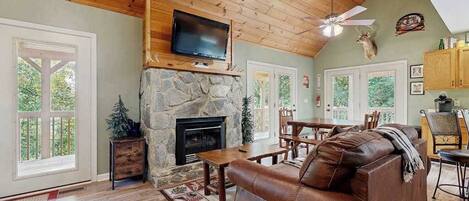 A cozy living room with a stone fireplace, leather sofa, wooden dining table, and a television mounted above the fireplace.