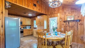 A wooden cabin interior with a dining table, chairs, a refrigerator, wooden cabinets, and a ceiling fan. - The Outpost (Whittier)