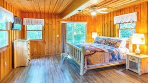 A wooden cabin bedroom with a bed, bedside table, and a television on the wall. - The Outpost (Whittier)
