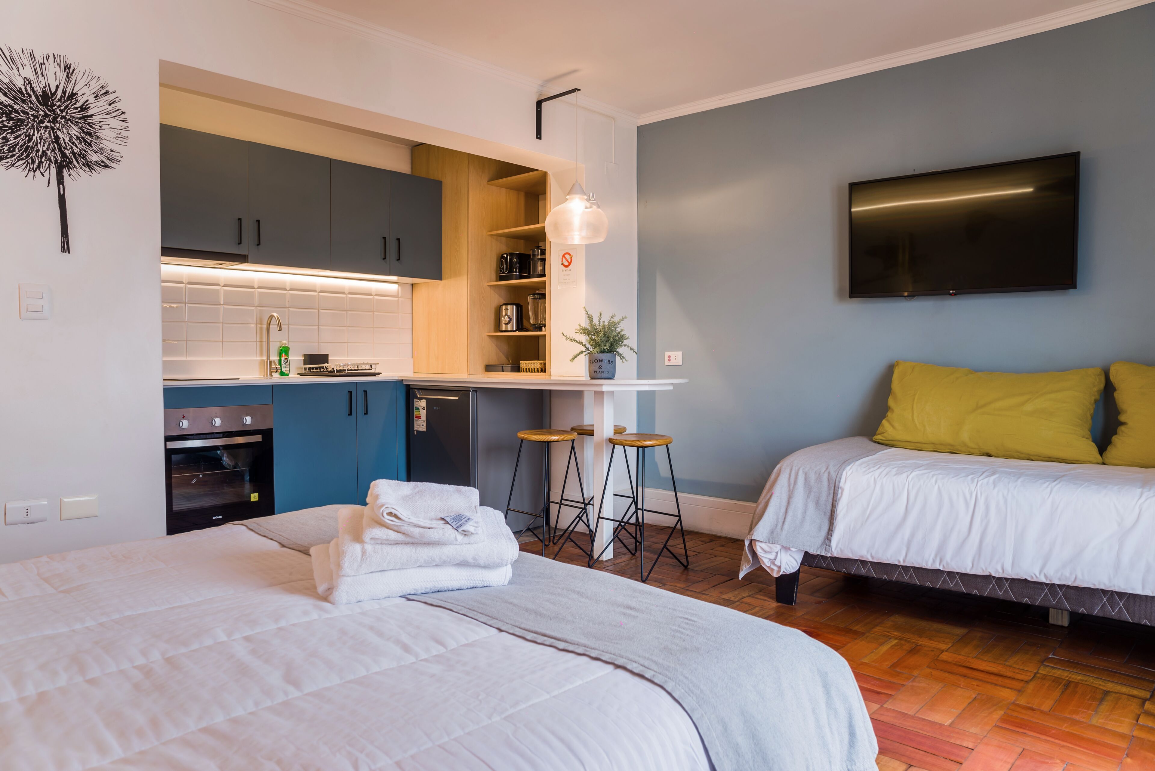 A modern hotel room with a kitchenette, a bed with white bedding, a TV mounted on the wall, and a wooden floor.