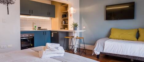 A modern hotel room with a kitchenette, a bed with white bedding, a TV mounted on the wall, and a wooden floor.