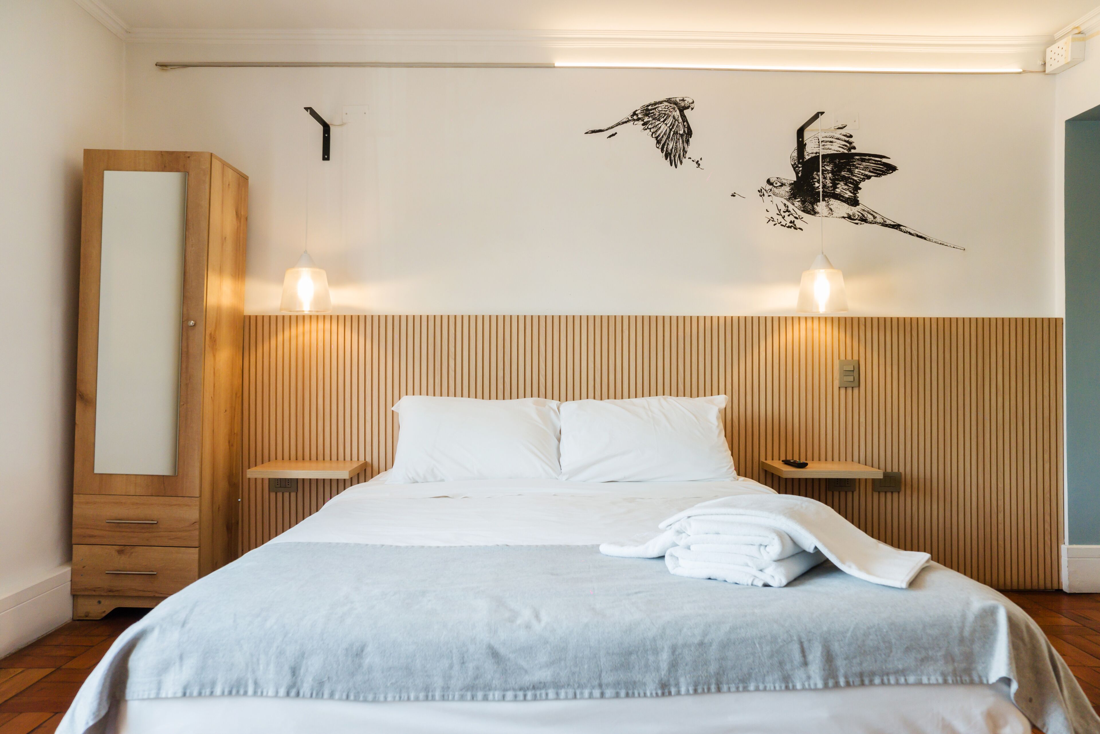 A hotel room with a bed, wooden headboard, two bedside lamps, a wall-mounted shelf, and a large mirror.