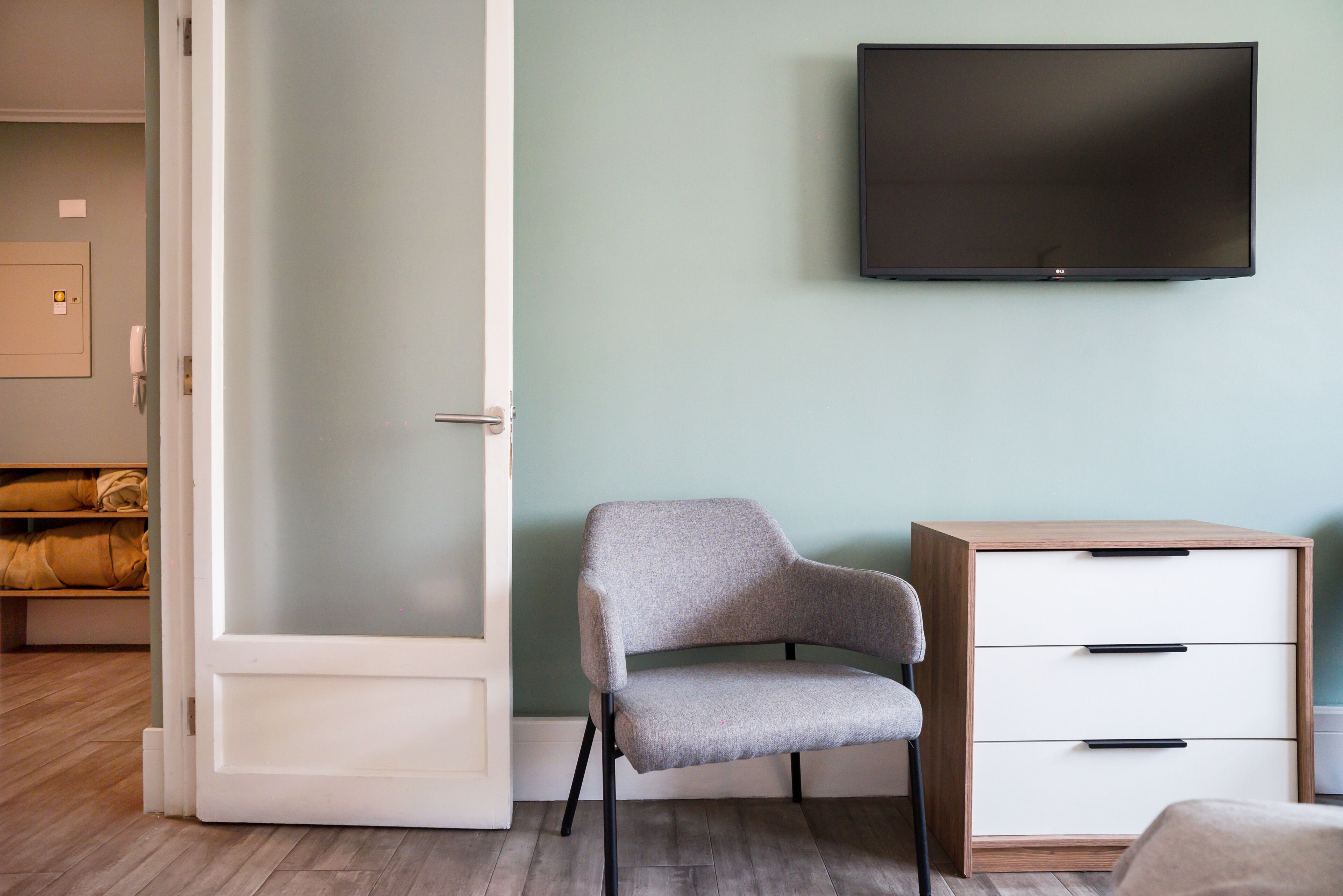 Ein Zimmer mit einem Sessel, einem Fernseher an der Wand, einer Holzkabine mit Schubladen und einer teilweise geöffneten Tür zu einem anderen Raum.