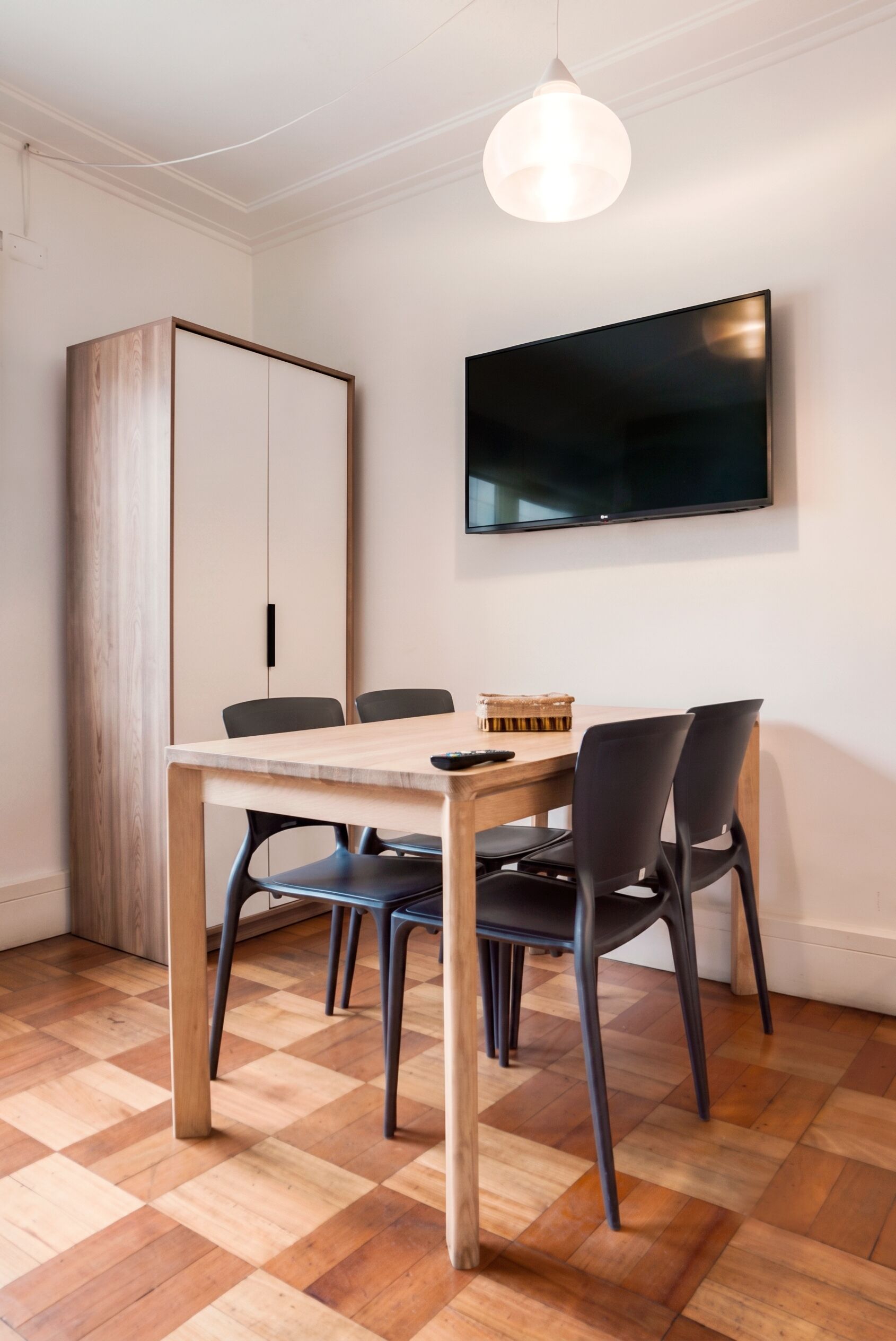 A dining area with a wooden table, black chairs, a television mounted on the wall, and a hanging light fixture.