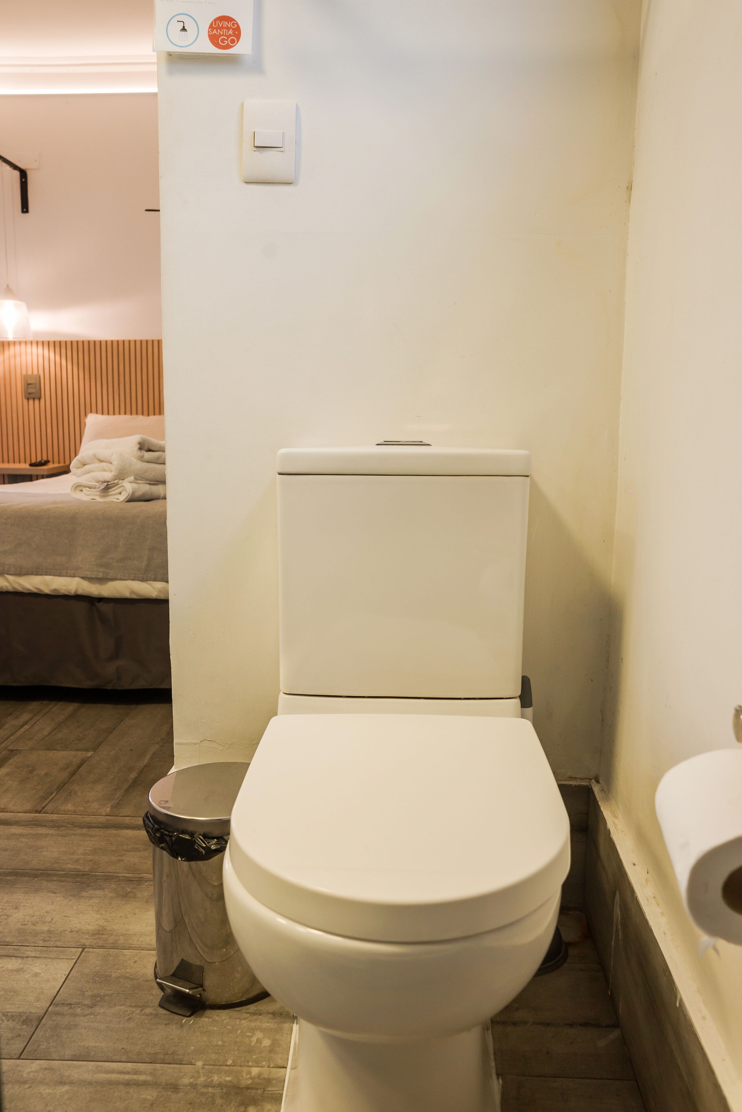 A bathroom with a toilet, a trash bin, and a bed with a wooden headboard in the background.