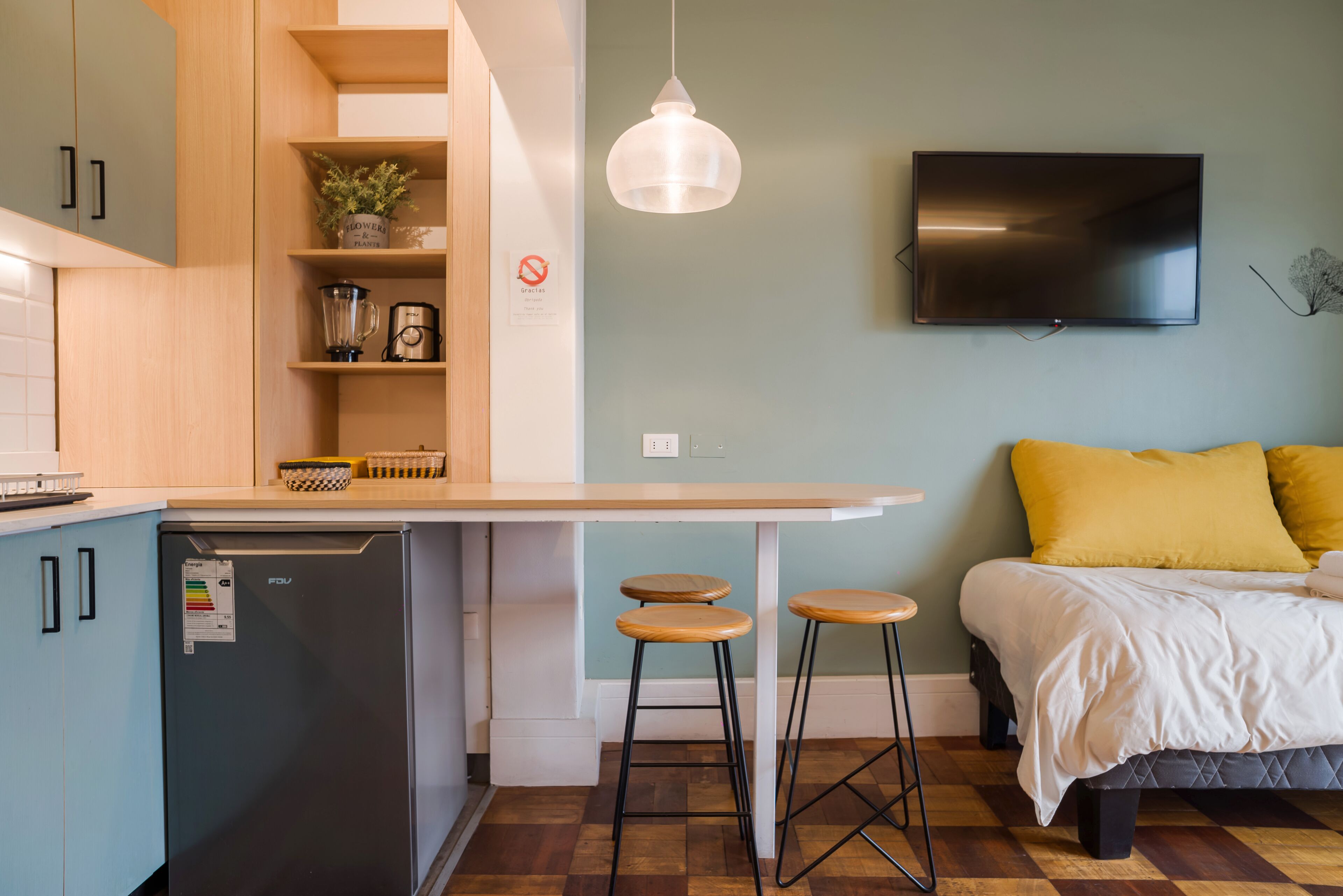 A compact living space with a kitchenette, a bed with yellow pillows, a TV mounted on the wall, and a bar stool.